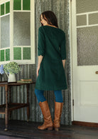 Woman standing backwards wearing a green corduroy dress and brown boots standing in a room with patterned wallpaper.