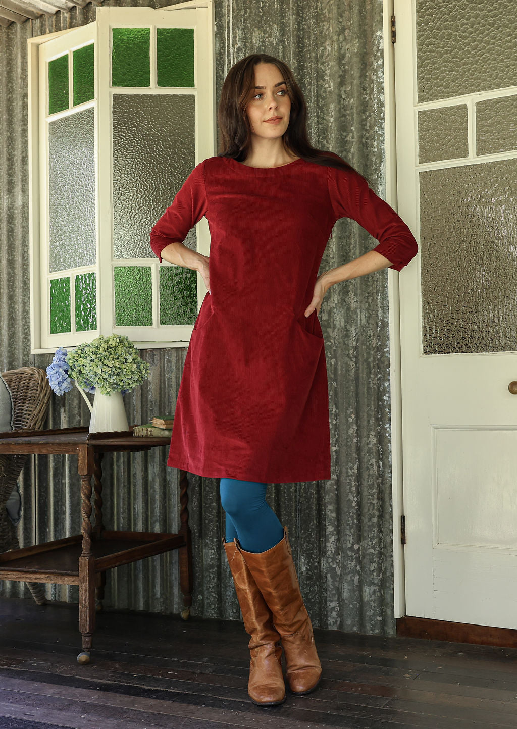 Woman in a red dress and brown boots standing in a room with patterned wallpaper.