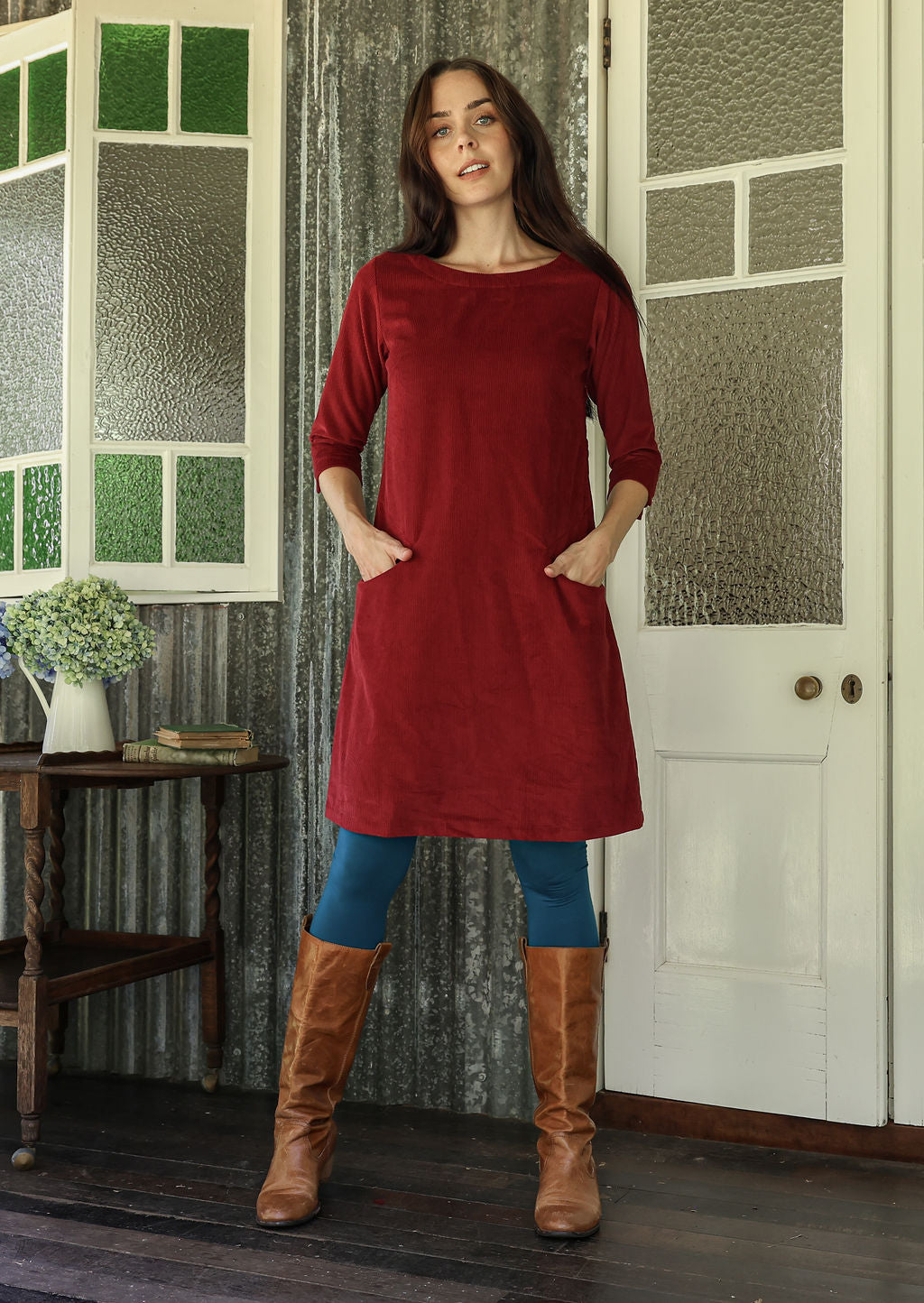 Woman wearing red corduroy A-line dress with 3/4 sleeves with hands in pockets over leggings and boots