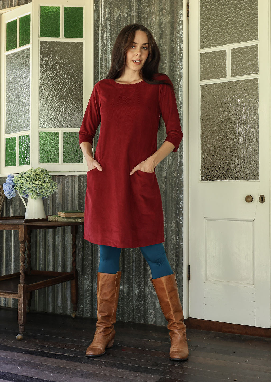 Woman wearing a red cotton corduroy dress and brown boots standing in a room with patterned walls.
