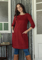 Woman wearing a red dress with blue leggings and brown boots standing in a room with patterned walls.