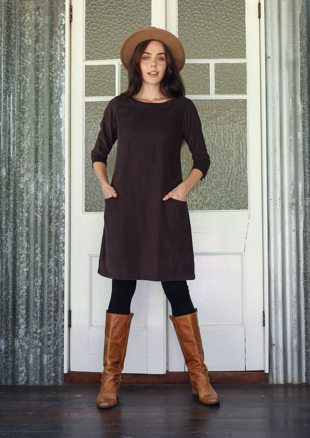 Woman wearing brown corduroy A-line dress with 3/4 sleeves with hands in pockets over leggings and boots