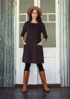 Woman wearing brown corduroy A-line dress with 3/4 sleeves with hands in pockets over leggings and boots