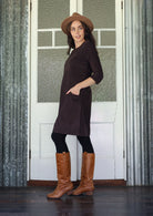 Woman standing sideways wearing a brown dress, hat, and boots standing in front of a white door with glass panels.