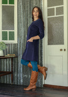 Woman standing sideways wearing a blue corduroy dress and brown boots standing in a room with patterned curtains and a door.