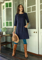 Woman in a navy blue corduroy dress with brown boots and a hat standing in a doorway.
