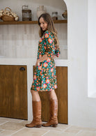 Woman standing sideways wearing a floral dress and brown boots standing in a kitchen setting.