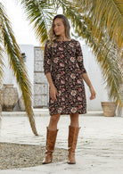 Woman wearing a black based floral print dress with brown boots standing against a rustic wooden door.