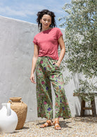 Model stands in sun wearing high waisted, relaxed fit cotton pants with drawstring and a hand in pocket. 