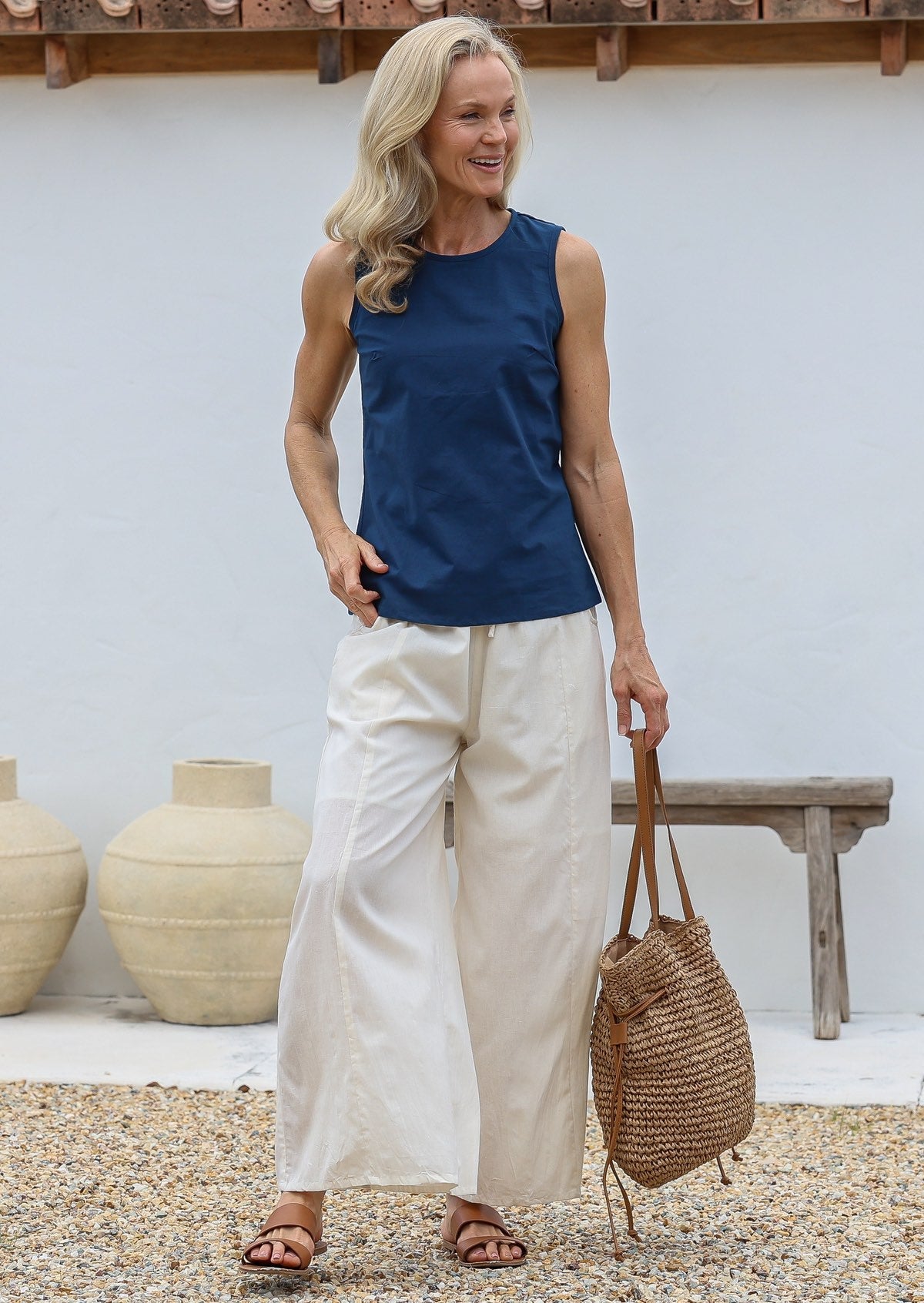 Woman wearing a blue sleeveless top and wide legged white cotton pants, holding a woven bag, standing against a white wall with decorative vases.