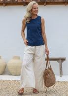 Woman wearing a blue sleeveless top and wide legged white cotton pants, holding a woven bag, standing against a white wall with decorative vases.