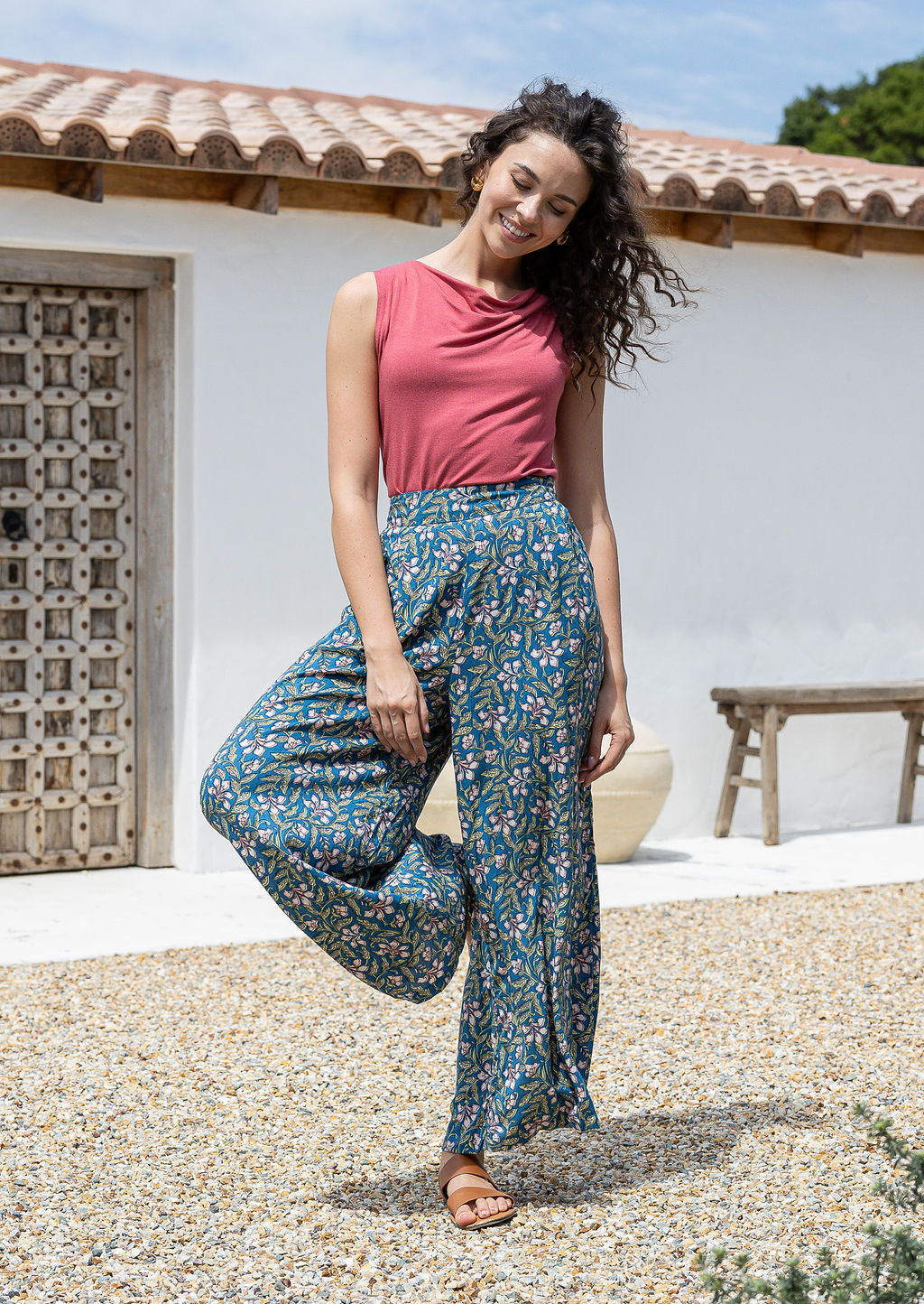 Woman wearing a pink top and rayon pants standing outdoors with a white wall and wooden door in the background.