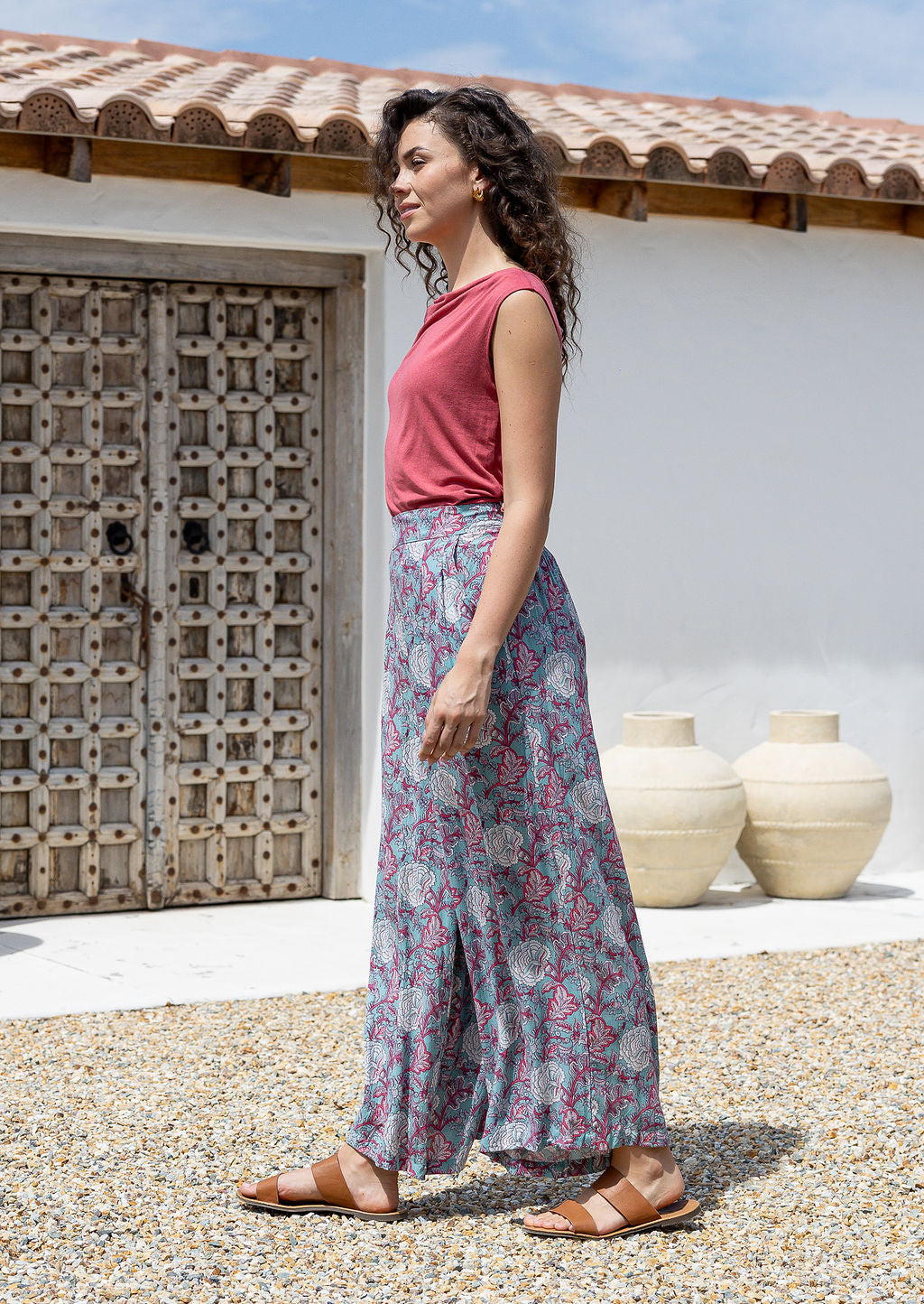 Woman wearing a pink top and rayon pants standing outdoors with a white wall and wooden door in the background.