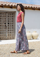 Woman wearing a pink top and rayon pants standing outdoors with a white wall and wooden door in the background.