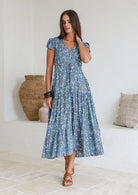 Woman poses in blue based block style floral print rayon maxi dress with V-neckline, buttoned bodice, waist-tie and pockets
