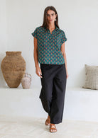Woman wearing a green based relaxed fit, shirt style floral cotton top with black plain pants.