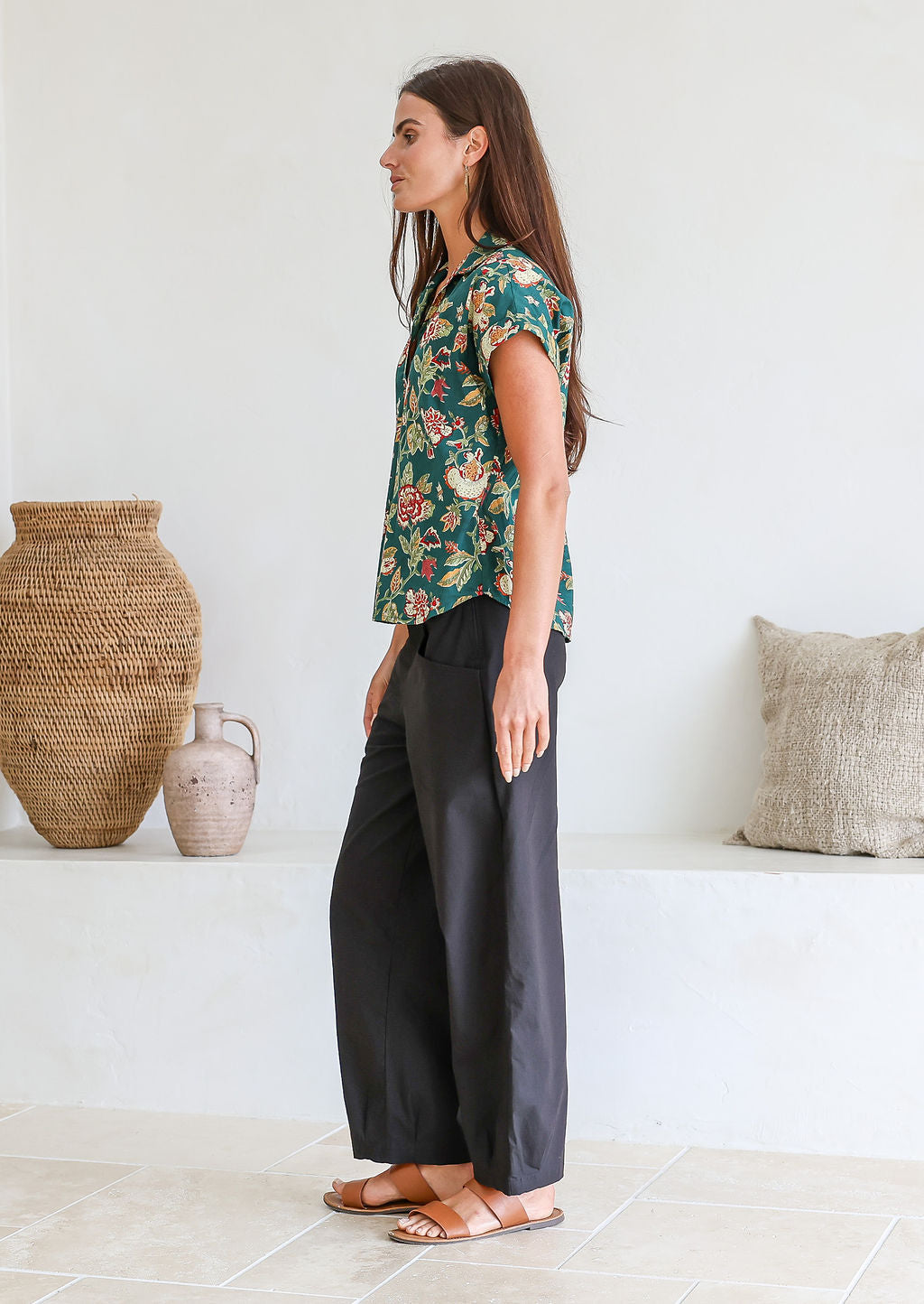Model stands sideways wearing a 100 percent cotton relaxed fit bottle green based floral top with short folded cuff sleeves, collar and V neckline