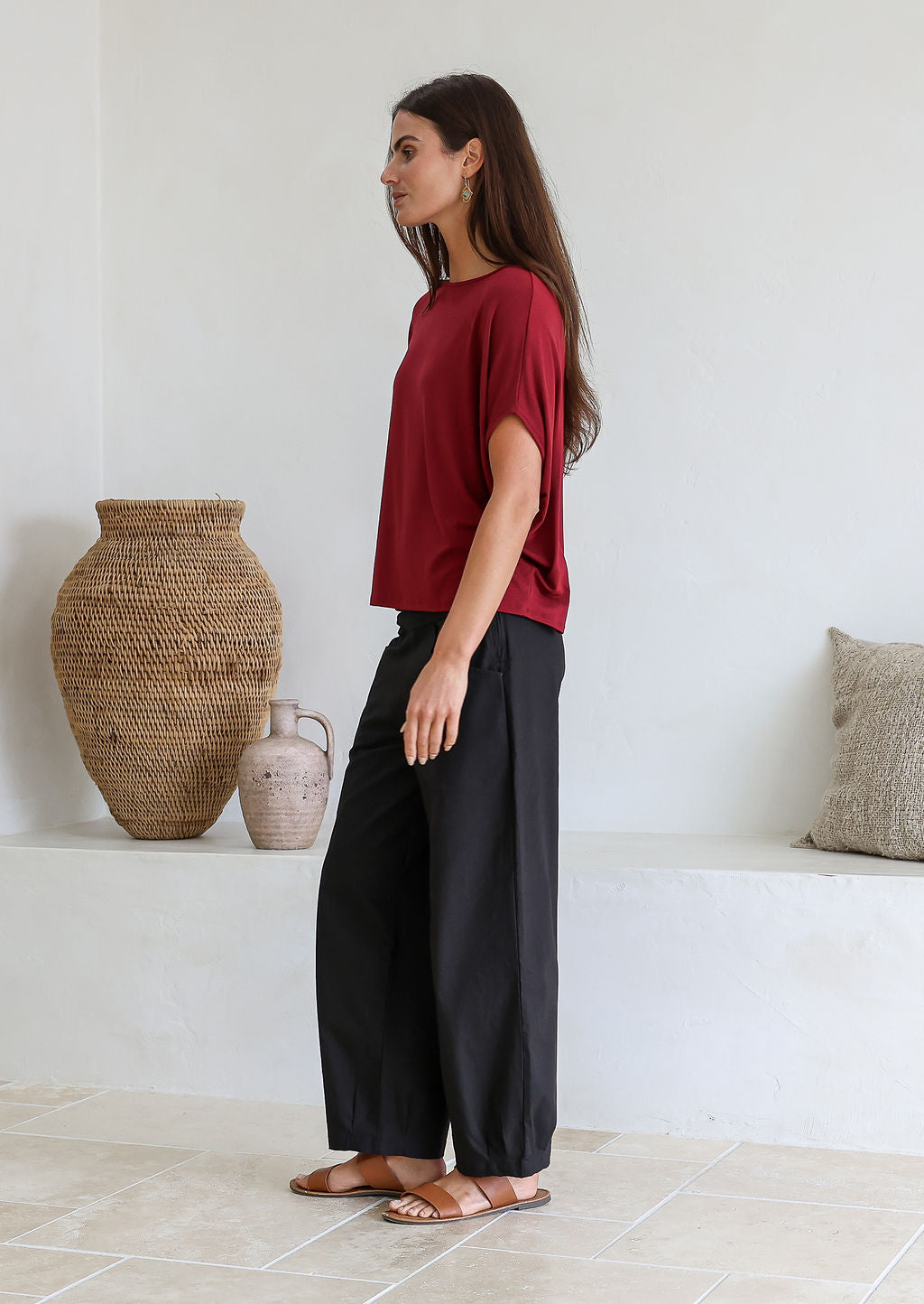 Woman standing sideways wearing a maroon relaxed fit stretch rayon t-shirt and black pants against a white wall.