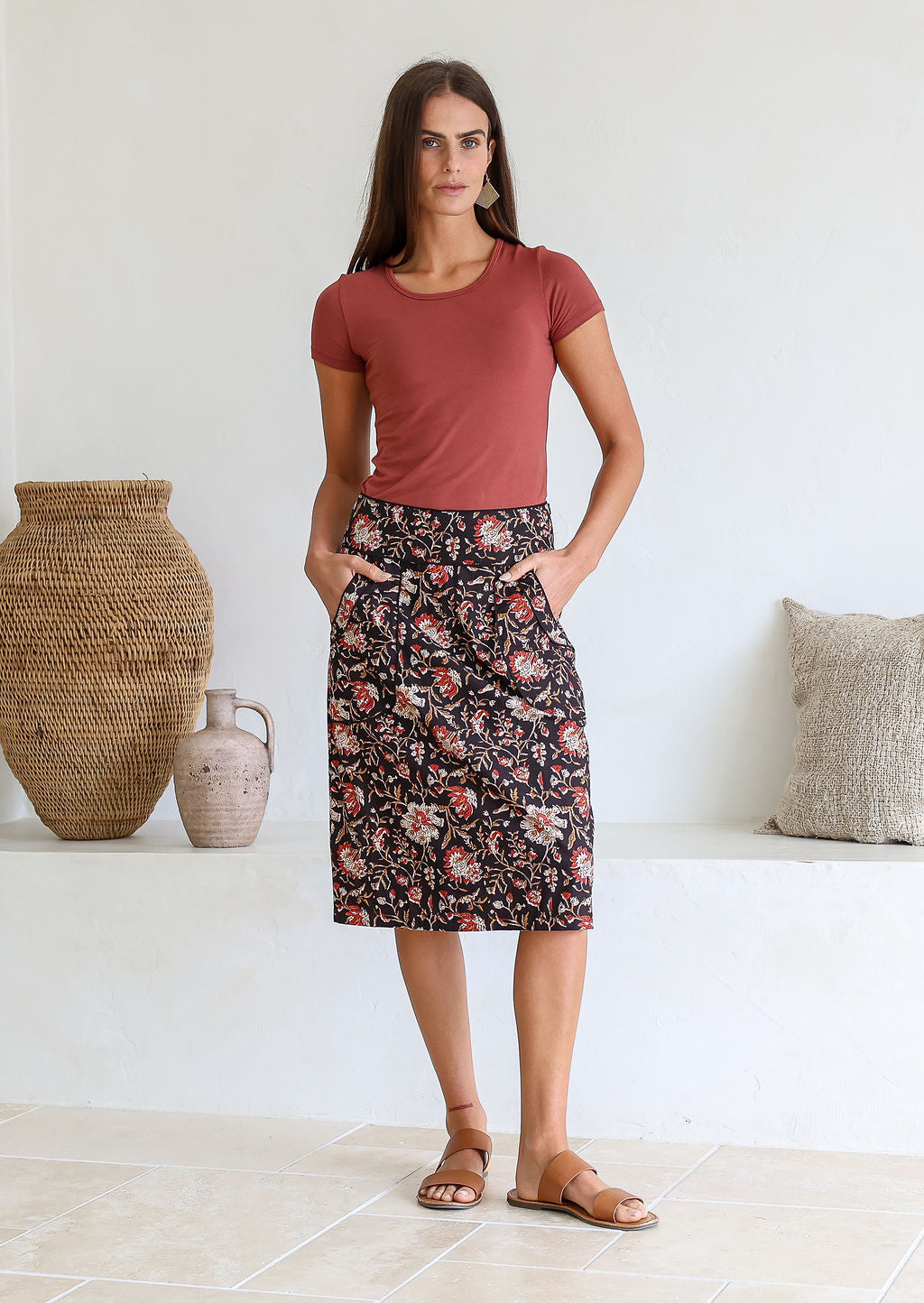 Woman wearing a pink top and black based floral print 100% cotton skirt standing in a room with decorative vases and pillows.