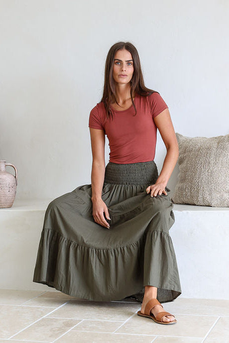 Model sitting on bench wearing a plain, cotton tiered maxi skirt with wide elasticated waistband.