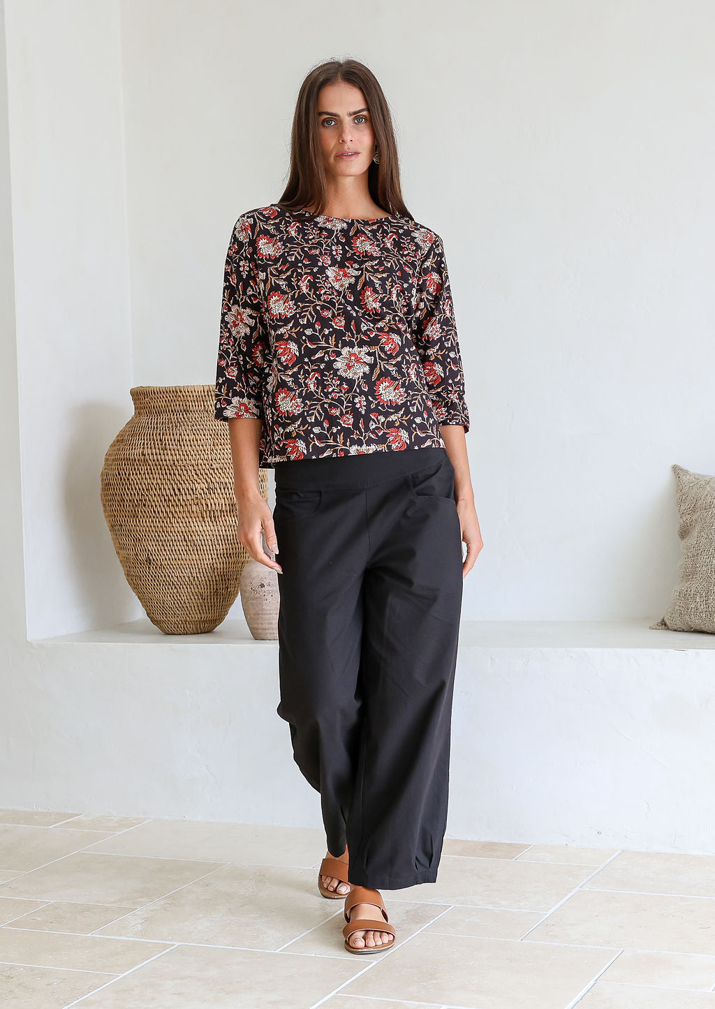 Woman wearing a black based floral top and black pants standing in a room with decorative vases