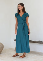 Woman wearing a teal rayon wrap maxi dress standing in a minimal indoor setting.