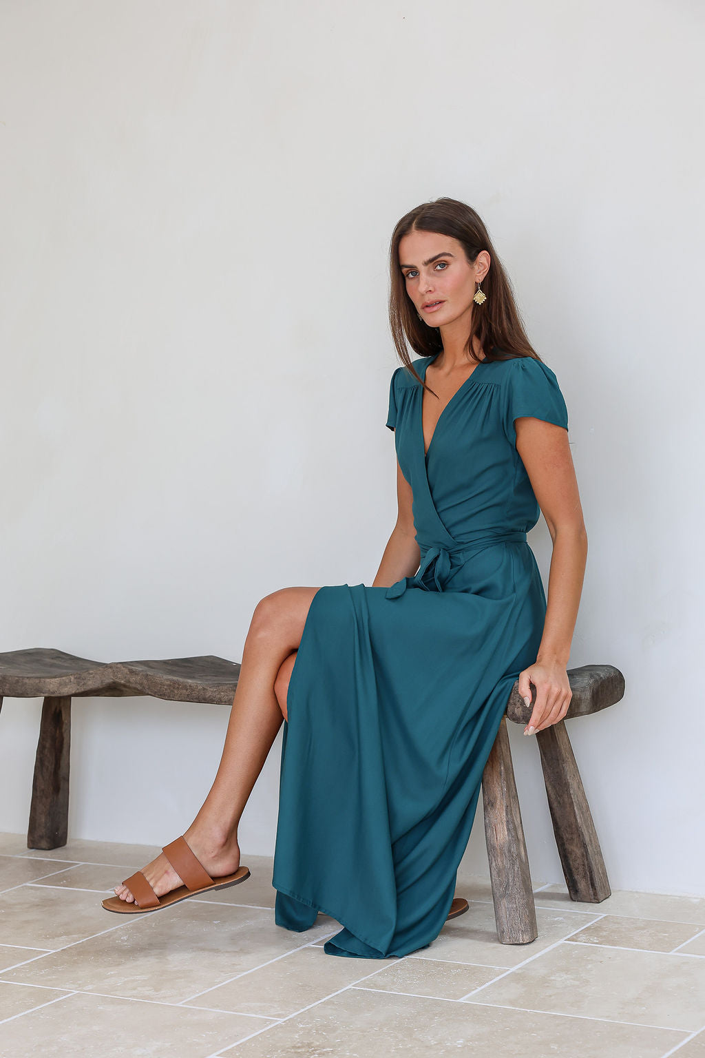 Woman in a teal rayon wrap dress sitting on a wooden stool against a white wall.