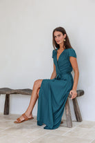Woman in a teal rayon wrap dress sitting on a wooden stool against a white wall.