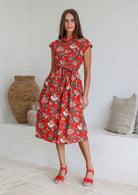 Model stands with wearing a midi length floral printed shirt style cotton dress with cap sleeves, buttoned bodice and a removable waist tie