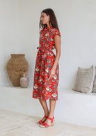 Model stands sideways near wearing a mid length, shirt style floral print cotton dress with blue, teal, green, pink, orange and tan on a on a red base.