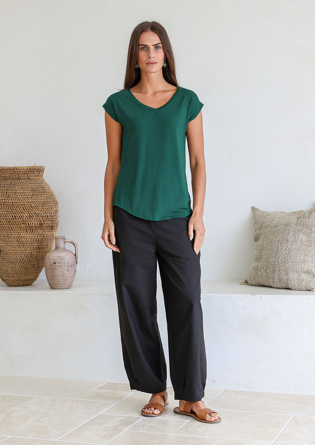 Woman wearing a 100% rayon forest green V-neck T-shirt and black pants, standing against a wall.