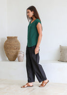 Woman stands sideways wearing a 100% rayon forest green V-neck top and black cotton pants standing against a white wall.