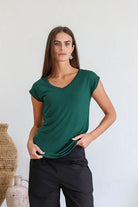 Woman wearing a 100% rayon forest green V-neck t-shirt and black pants standing against a white wall.