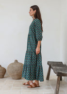 Woman standing in front of wooden bench wears three tiered cotton maxi dress with 3/4 sleeves, pockets and mandarin collar