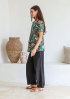 Woman stands sideways wearing a relaxed fit bottle green based floral print top against a white wall.