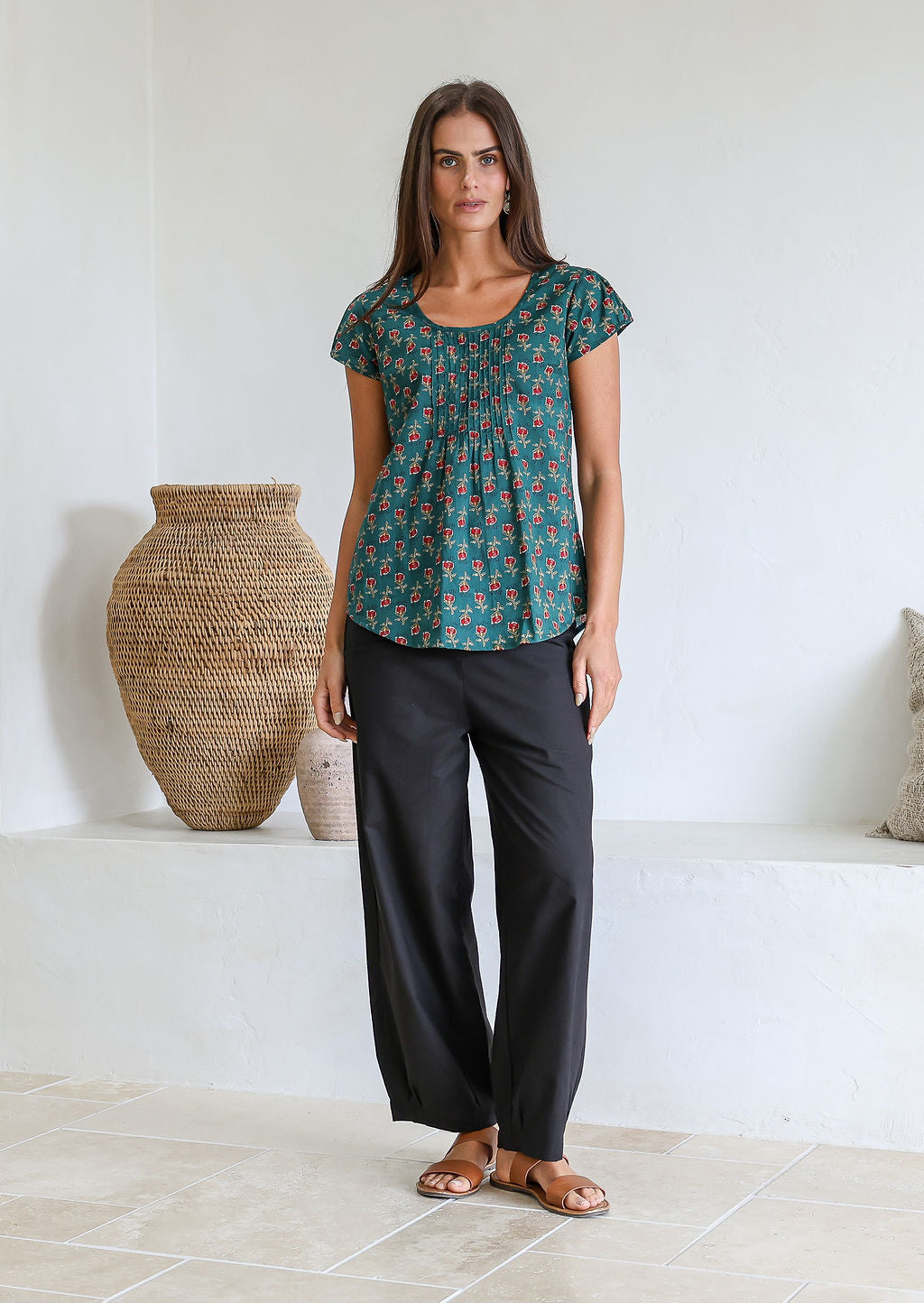 Woman wears bottle green based floral 100t% cotton cap sleeve top with pin tuck detail across the bust and a scoop neckline