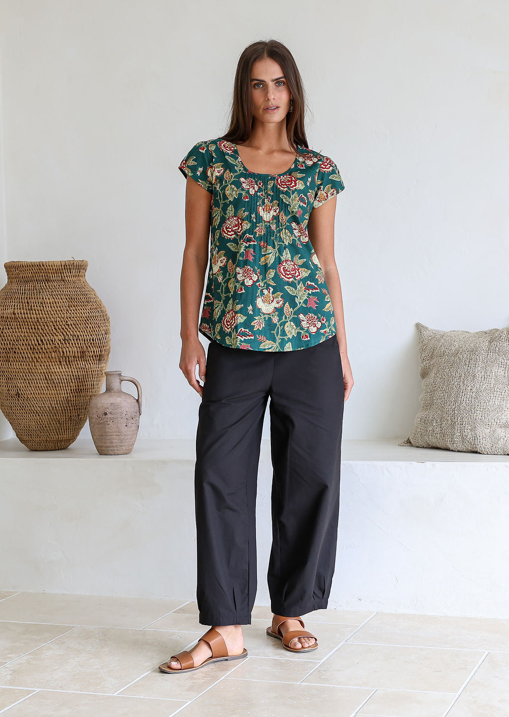 Woman wears bottle green based floral print 100% cotton cap sleeve top with pin tuck detail across the bust and a scoop neckline