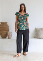 Woman wears bottle green based floral print 100% cotton cap sleeve top with pin tuck detail across the bust and a scoop neckline