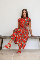 Woman in a red based floral print 100% cotton dress sitting on a wooden bench against a white wall.