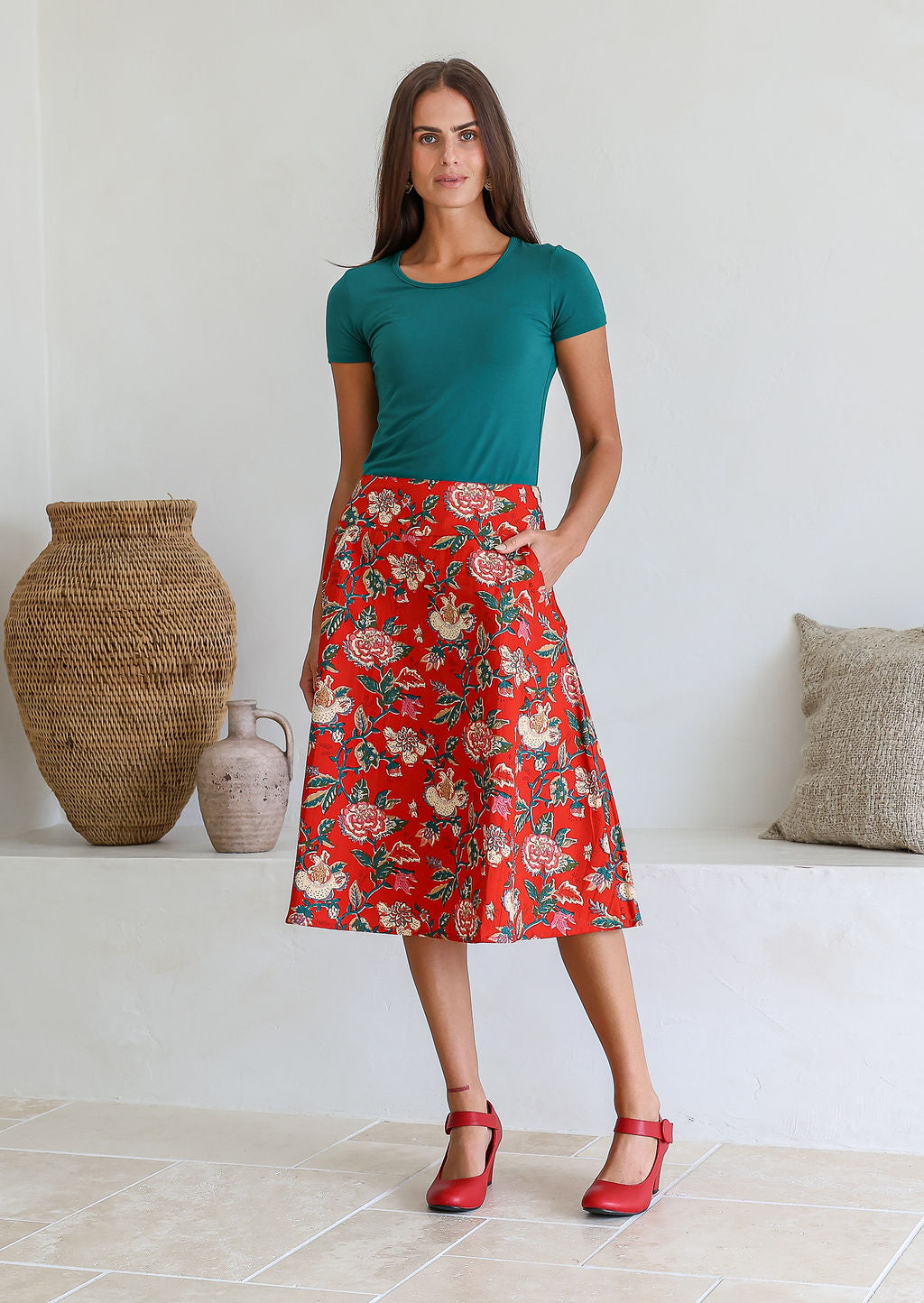 Woman wearing a basic green top and red floral a-line cotton skirt with side zipper and one hand in pocket.