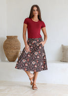 Woman wearing a maroon top and floral  cotton skirt standing in a room with a woven vase and pillows.