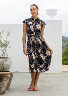 Woman wearing a midi length black based floral cotton dress with buttoned bodice, standing against a white wall and decorative vases