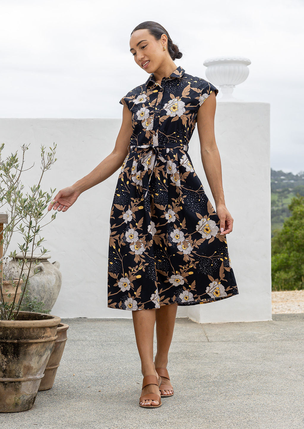 Model stands with wearing a midi length floral printed shirt style cotton dress with cap sleeves, buttoned bodice and a removable waist tie