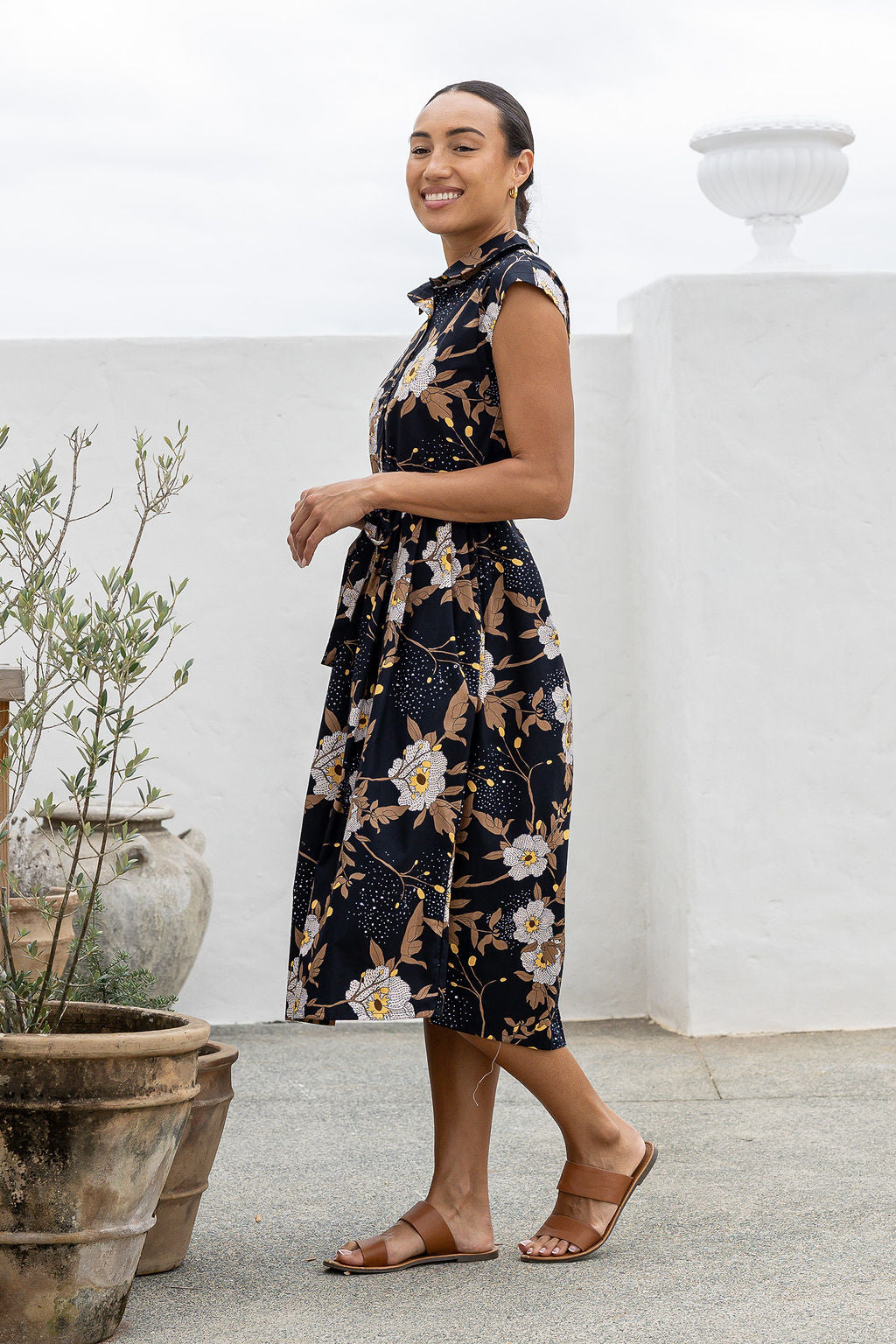Model stands sideways near wearing a mid length, shirt style cotton dress with black based floral print