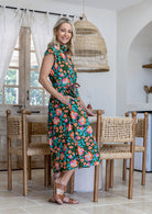 Model stands wearing a mid length, shirt style floral print cotton dress in olive green based floral print with one hand in pocket