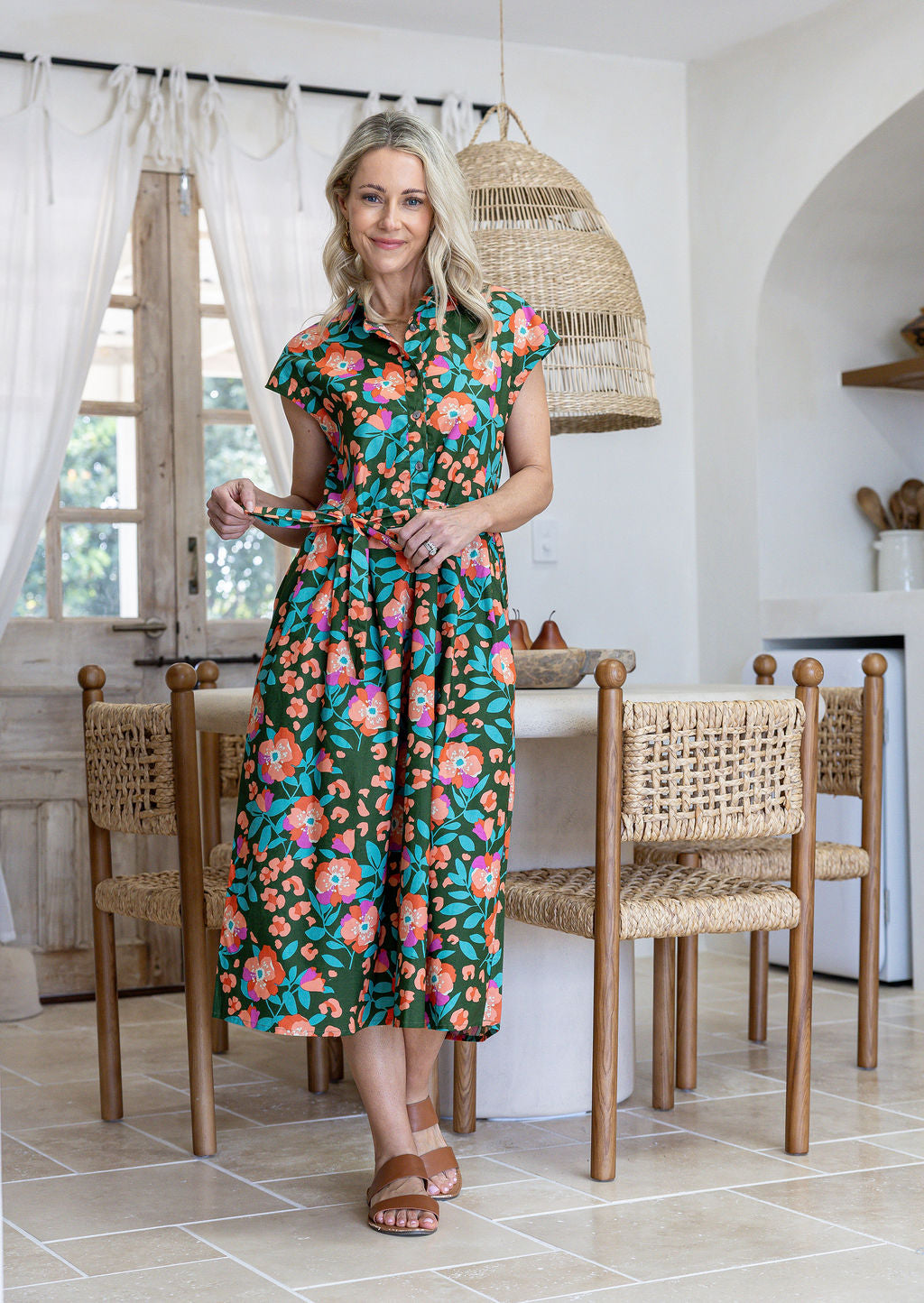 Woman wearing a midi length olive green based floral cotton dress with buttoned bodice and a removable waist tie.