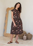 Model stands with wearing a midi length floral printed shirt style cotton dress with cap sleeves, buttoned bodice and a removable waist tie