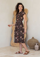 Model stands sideways near ceramic pots, wearing a midlength, shirt style Indian traditional  floral print cotton dress with with rust, tan and sand tones on a black base.