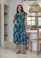 A woman wearing a cap-sleeved, collared cotton midi dress with a multicolour peacock and floral print on a dark navy base