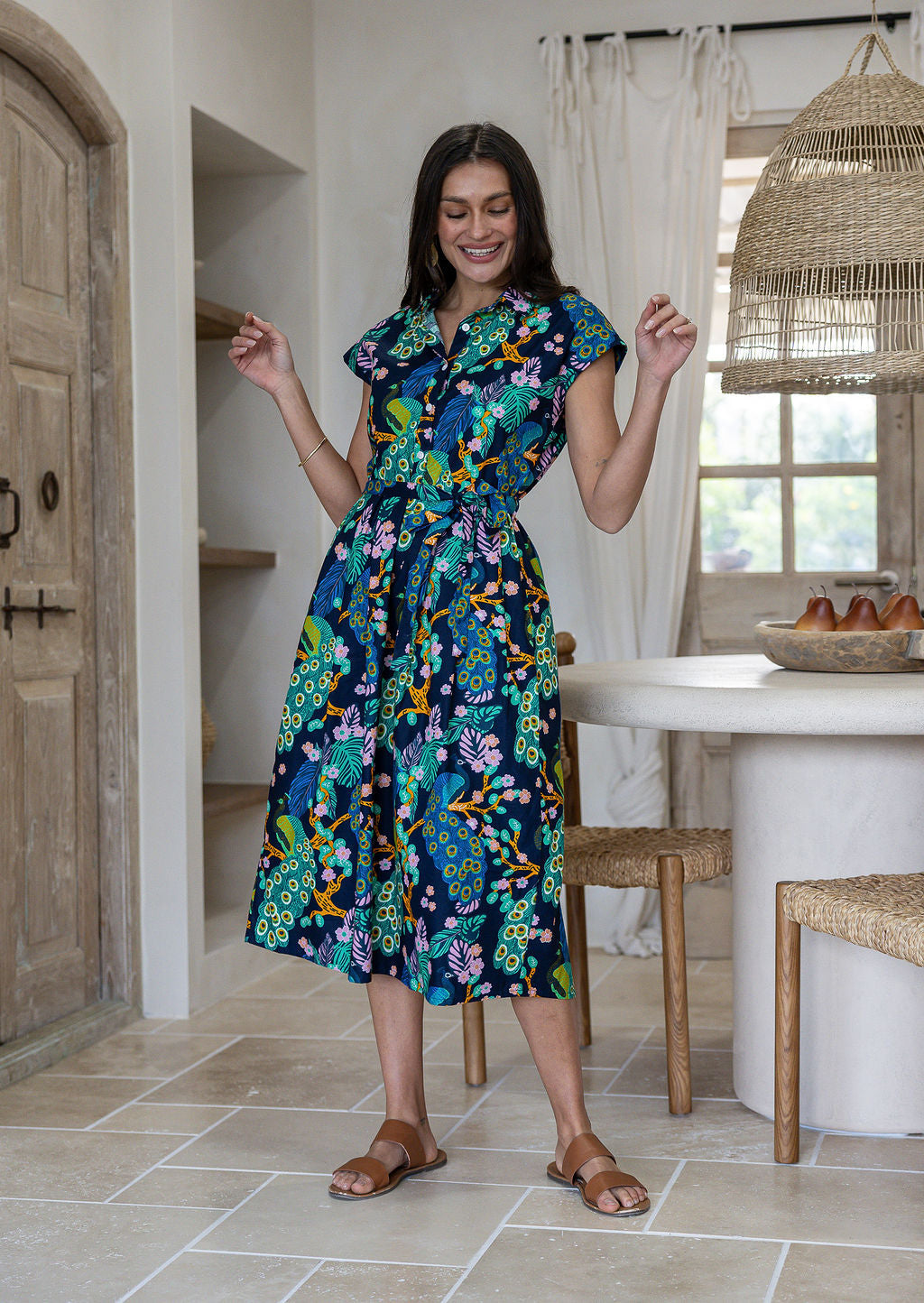 A woman modelling a cap-sleeve cotton midi dress with a vibrant floral and peacock print in teal, green, blue, yellow, and pink on a dark navy background, set in a bright, modern rustic kitchen.